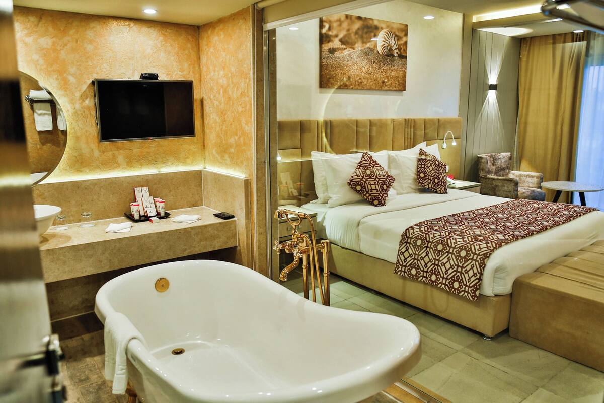 A modern hotel room with a large bed, a bathtub, a TV mounted on the wall, and a bathroom with a sink and mirror.