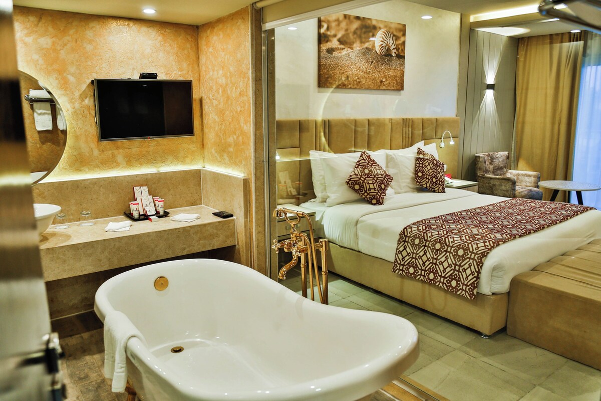 A modern hotel room with a large bed, a bathtub, a TV mounted on the wall, and a bathroom with a sink and mirror.