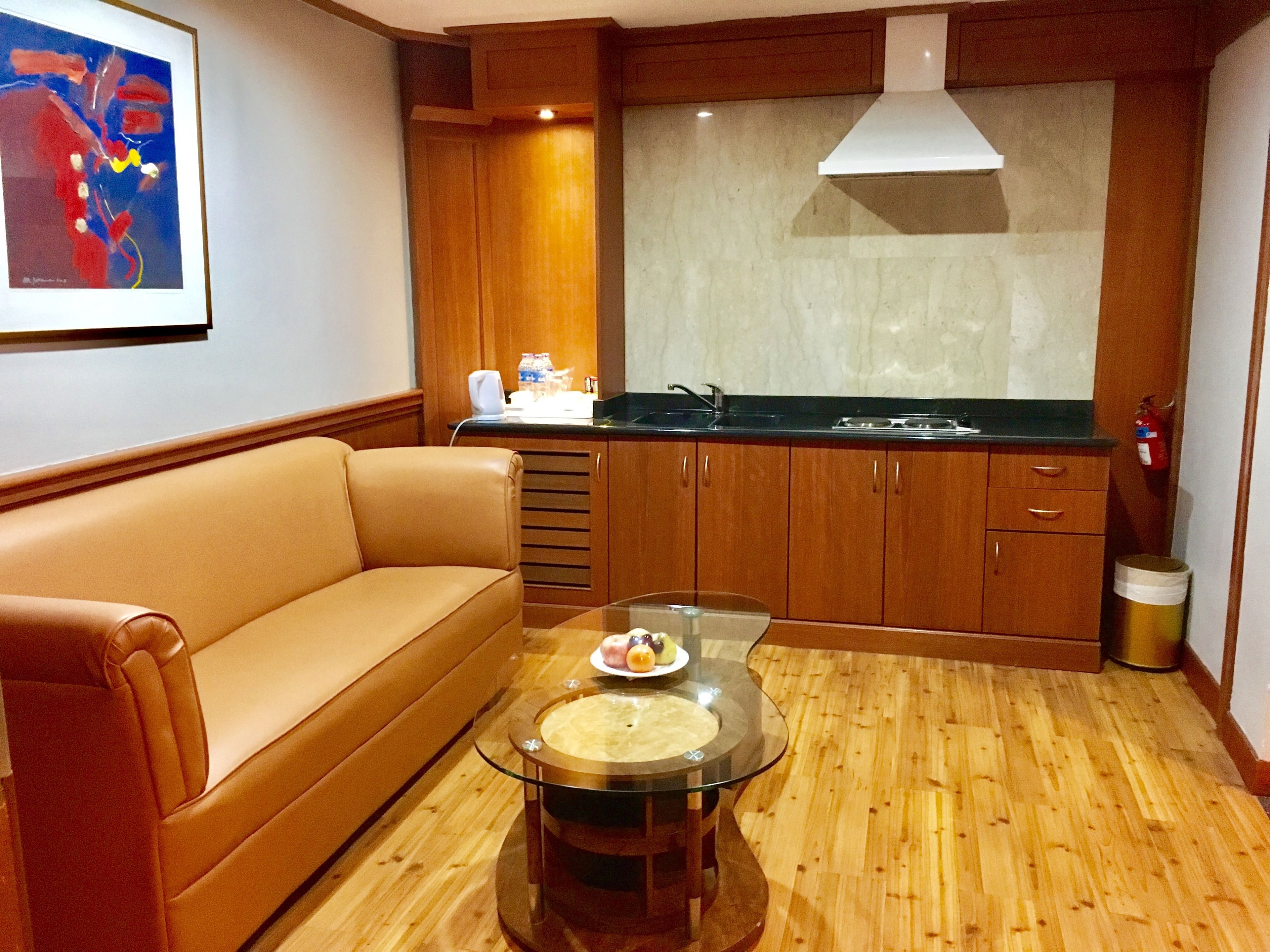A compact living space with a sofa, a glass-top table with a bowl, wooden cabinets, a kitchen area with a sink, and a framed abstract painting on the wall.