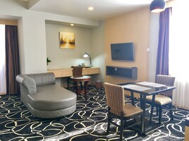 Habitación de hotel con sofá, una mesa de comedor con sillas, un escritorio con lámpara, televisor y alfombra estampada.