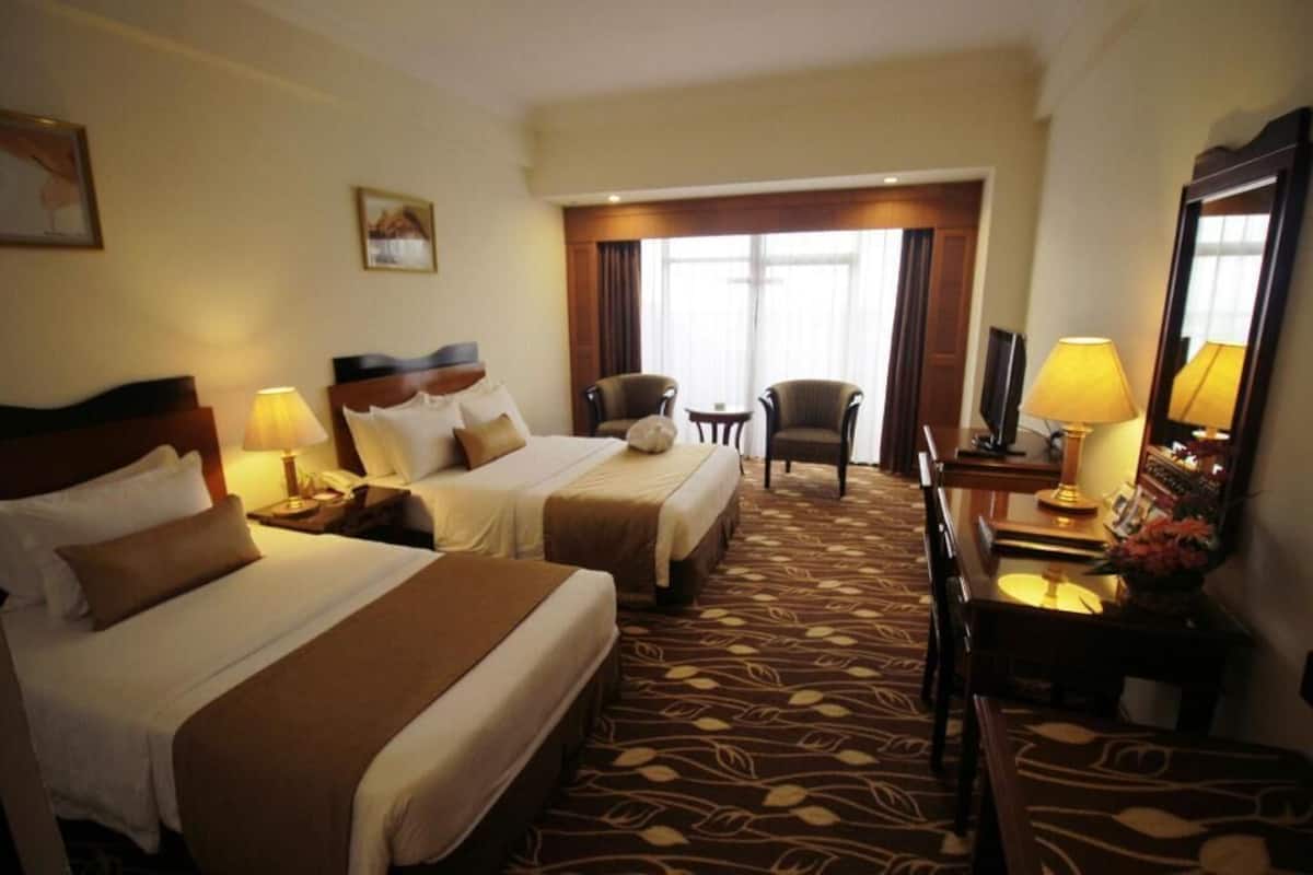 A hotel room with two beds, a desk with a lamp, a chair, and a large window.