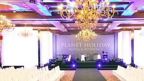 Escenario de hotel con un cartel que dice 'PLANET HOLIDAY HOTEL & RESIDENCE' y filas de sillas blancas mirando al escenario.