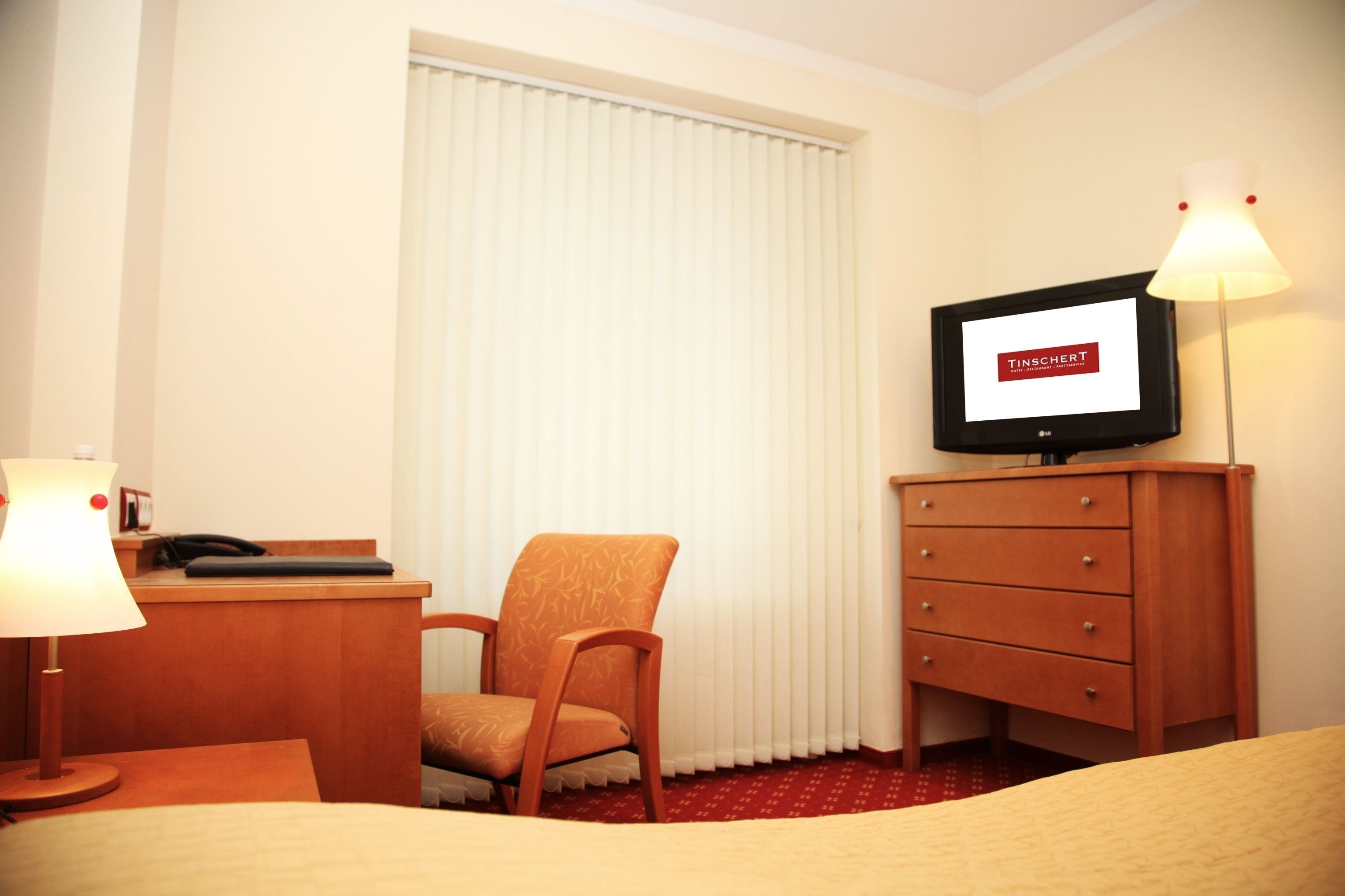 A hotel room with a bed, a desk with a lamp, a chair, a TV on a dresser, and a window with blinds.
