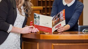 Two individuals at a reception desk with brochures, a clock, and a wooden panel wall in the background. - Tinschert Hotel-Restaurant-Partyservice (Schwertberg)