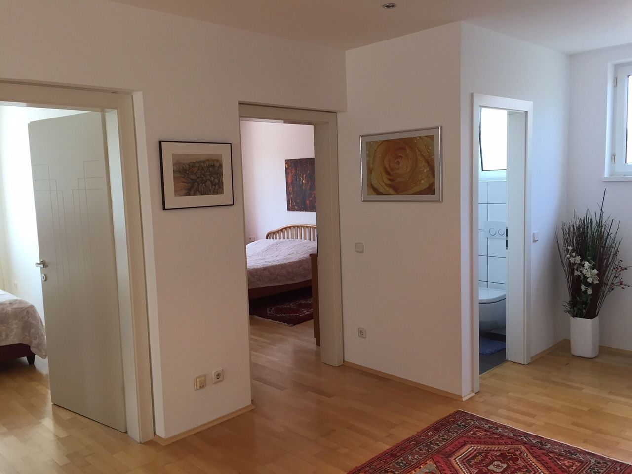 Amplia sala de estar con piso de madera, una cama y un tapete grande. Se observan obras de arte enmarcadas en las paredes y una planta en maceta en una esquina.