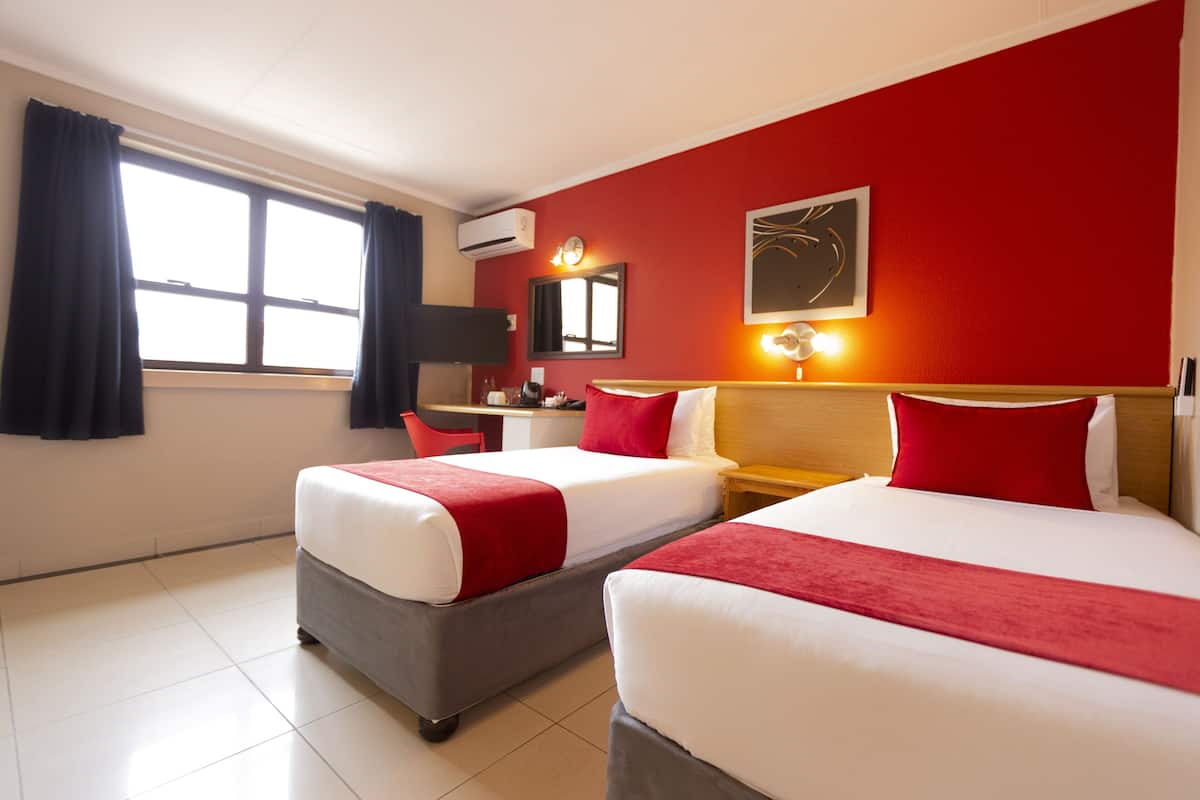 Quarto de hotel com duas camas, uma mesa, uma cadeira, televisão e janela com cortinas.