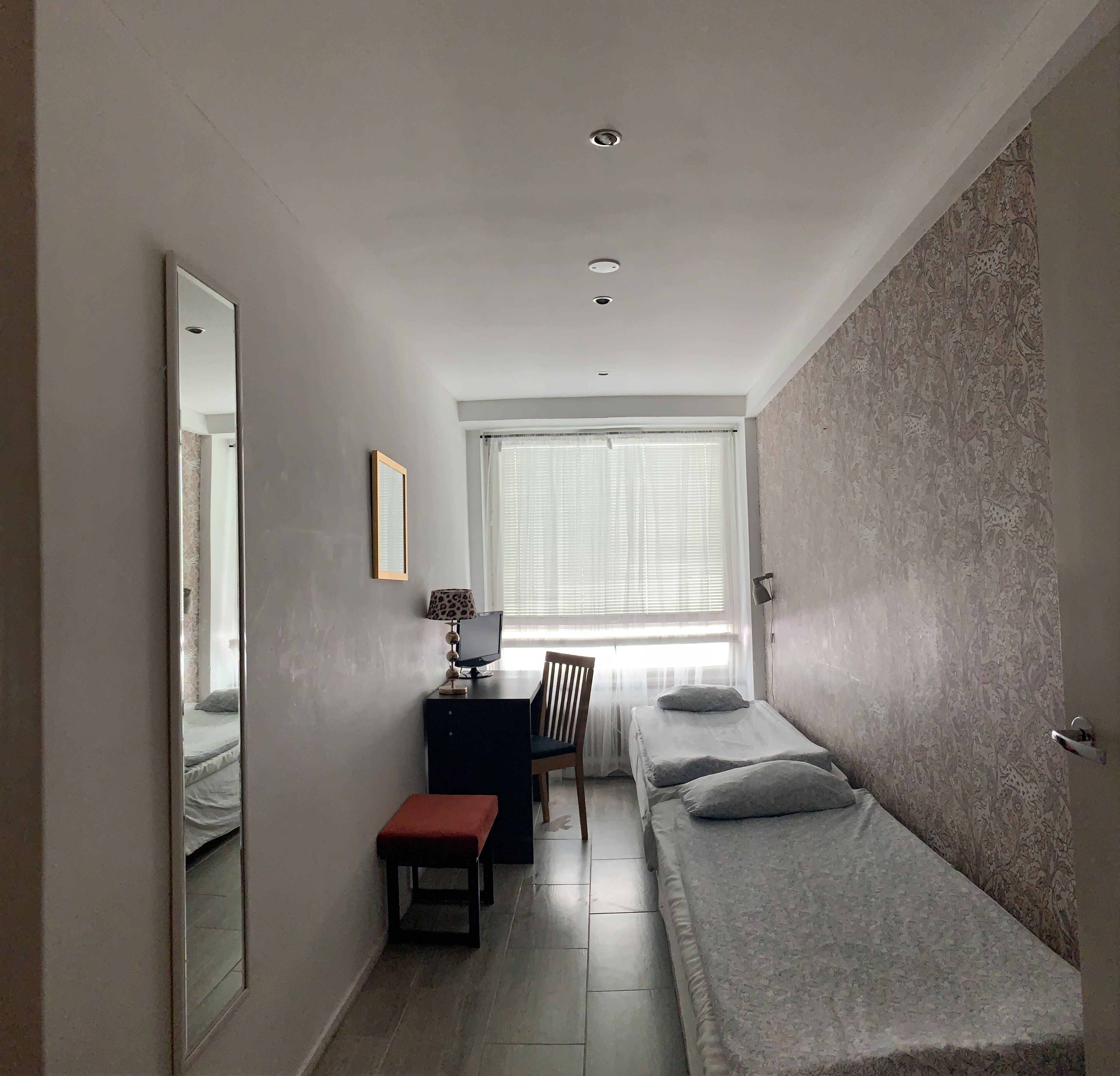 A small, well-lit room with a bed, a desk with a chair, a mirror, and a window with blinds.