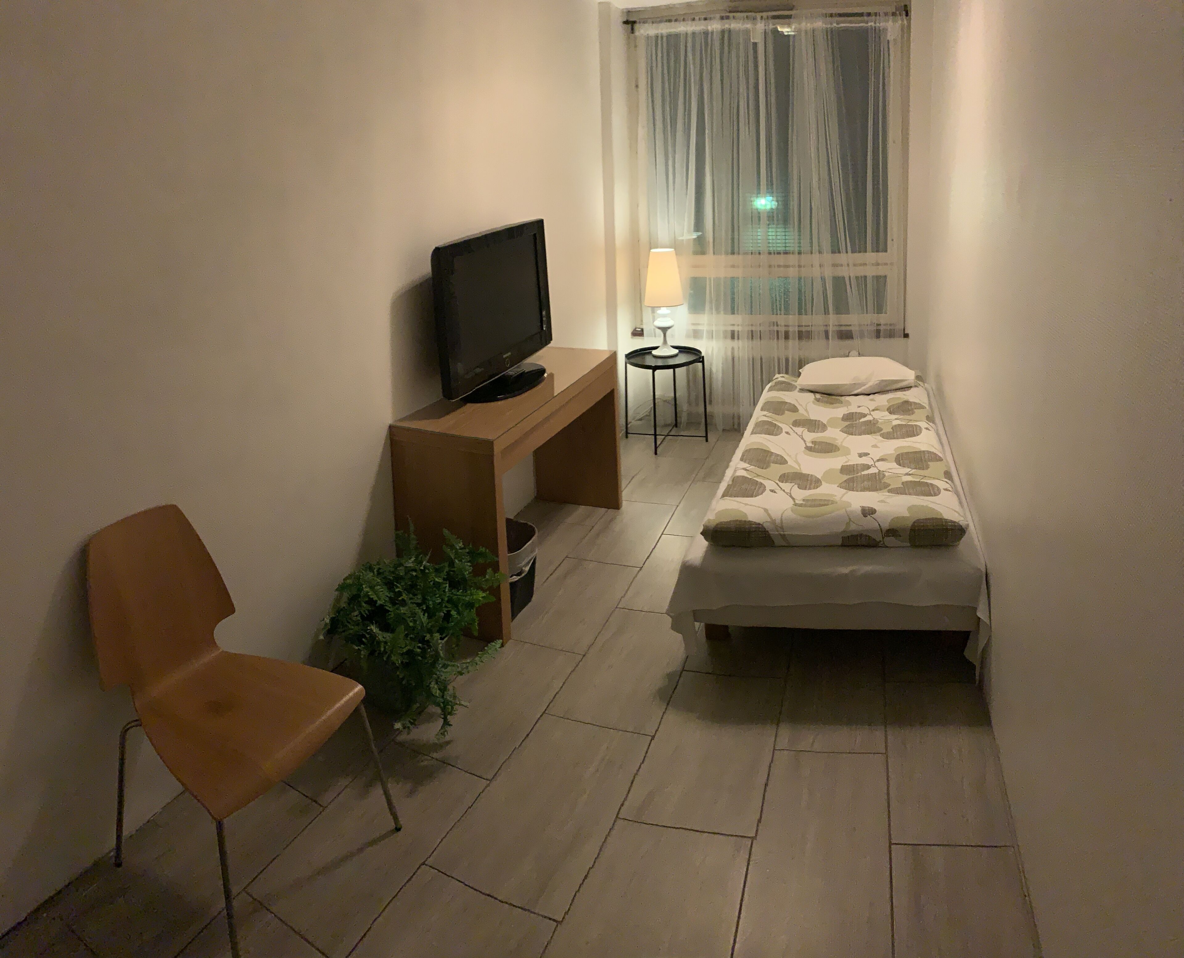 Une petite pièce étroite, avec un lit simple, un téléviseur posé sur un meuble en bois, une chaise, une petite table avec une lampe et une fenêtre ornée de rideaux légers.