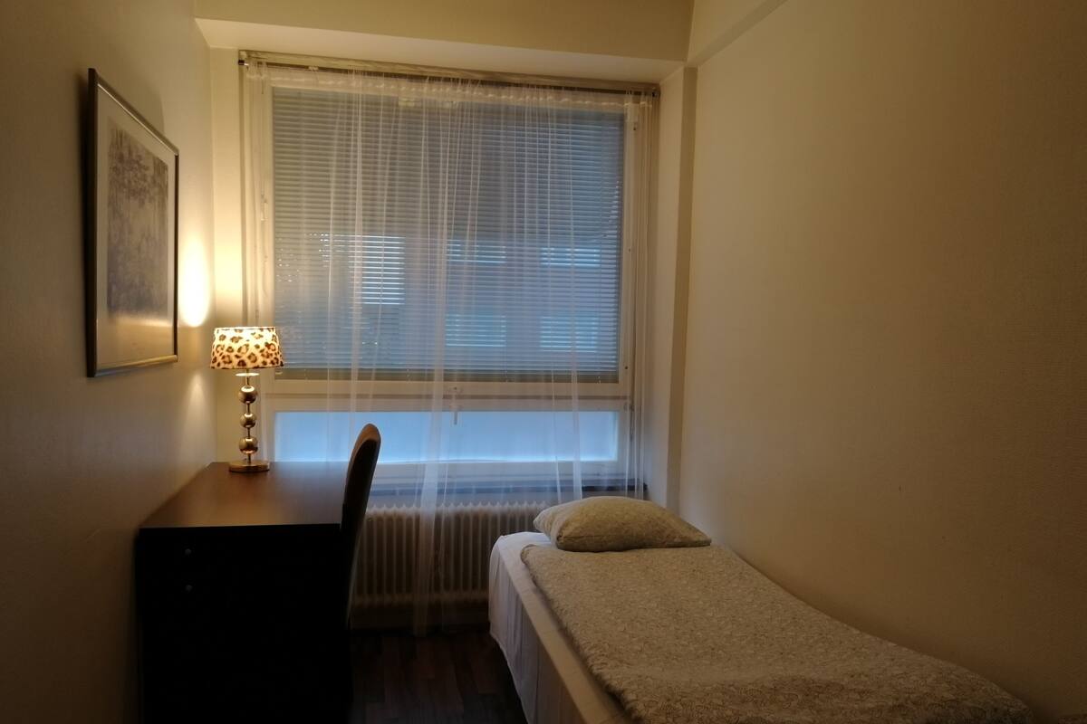 A small, dimly lit room with a single bed, a desk with a lamp, and a radiator under a window with blinds.