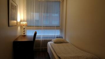A small, dimly lit room with a single bed, a desk with a lamp, and a radiator under a window with blinds.
