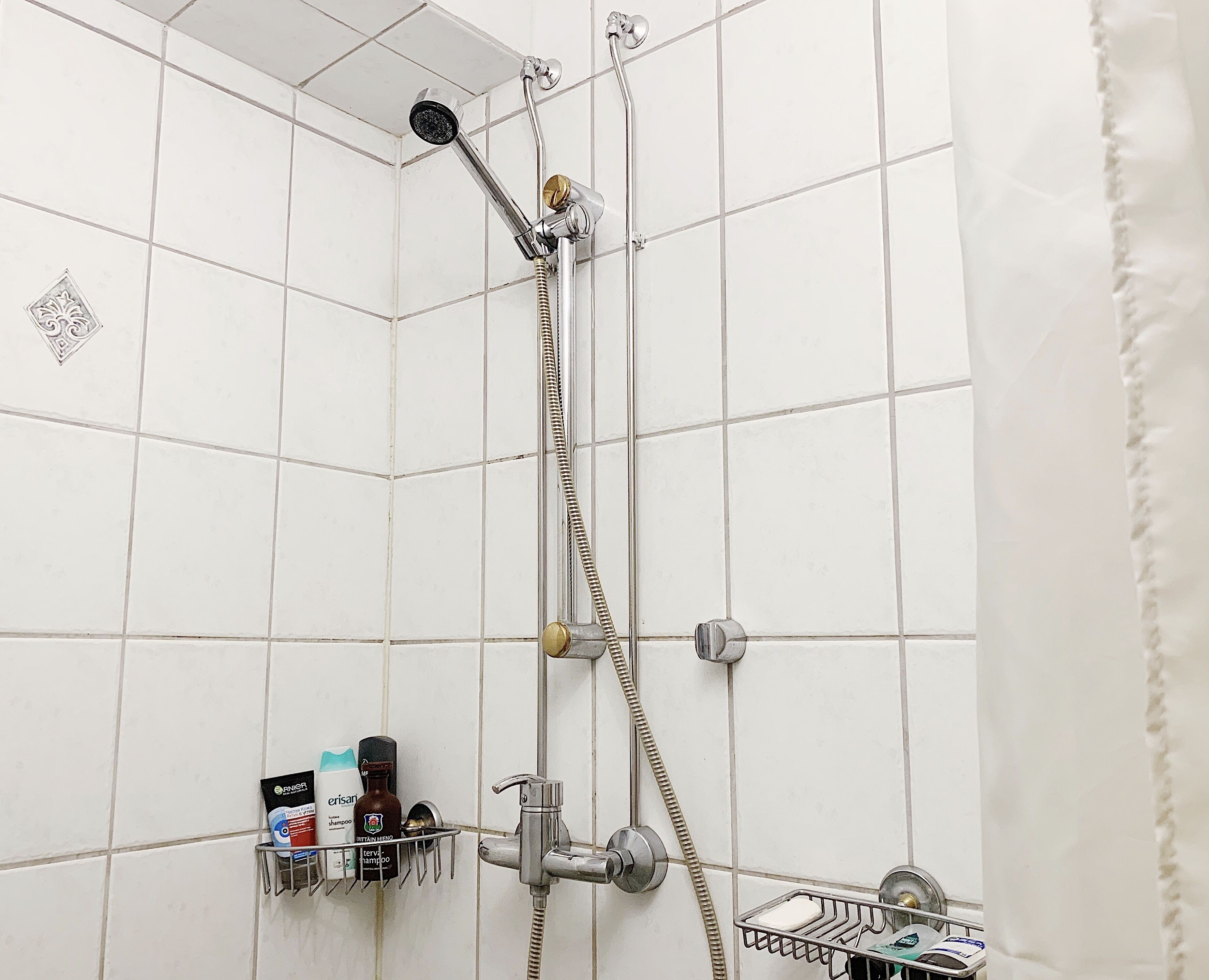 A shower with a handheld showerhead and a wall-mounted faucet. There are toiletries on a shelf, including a bottle of shampoo and a razor.