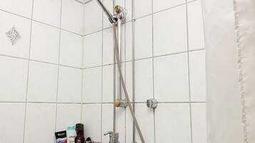 A shower with a handheld showerhead and a wall-mounted faucet. There are toiletries on a shelf, including a bottle of shampoo and a razor.