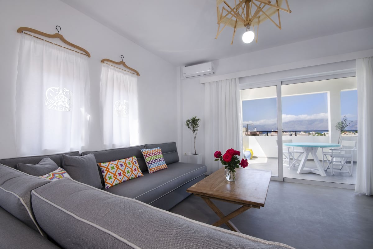 Un salón moderno con un sofá gris, almohadones coloridos, una mesa de centro de madera y vistas al mar a través de grandes ventanales.