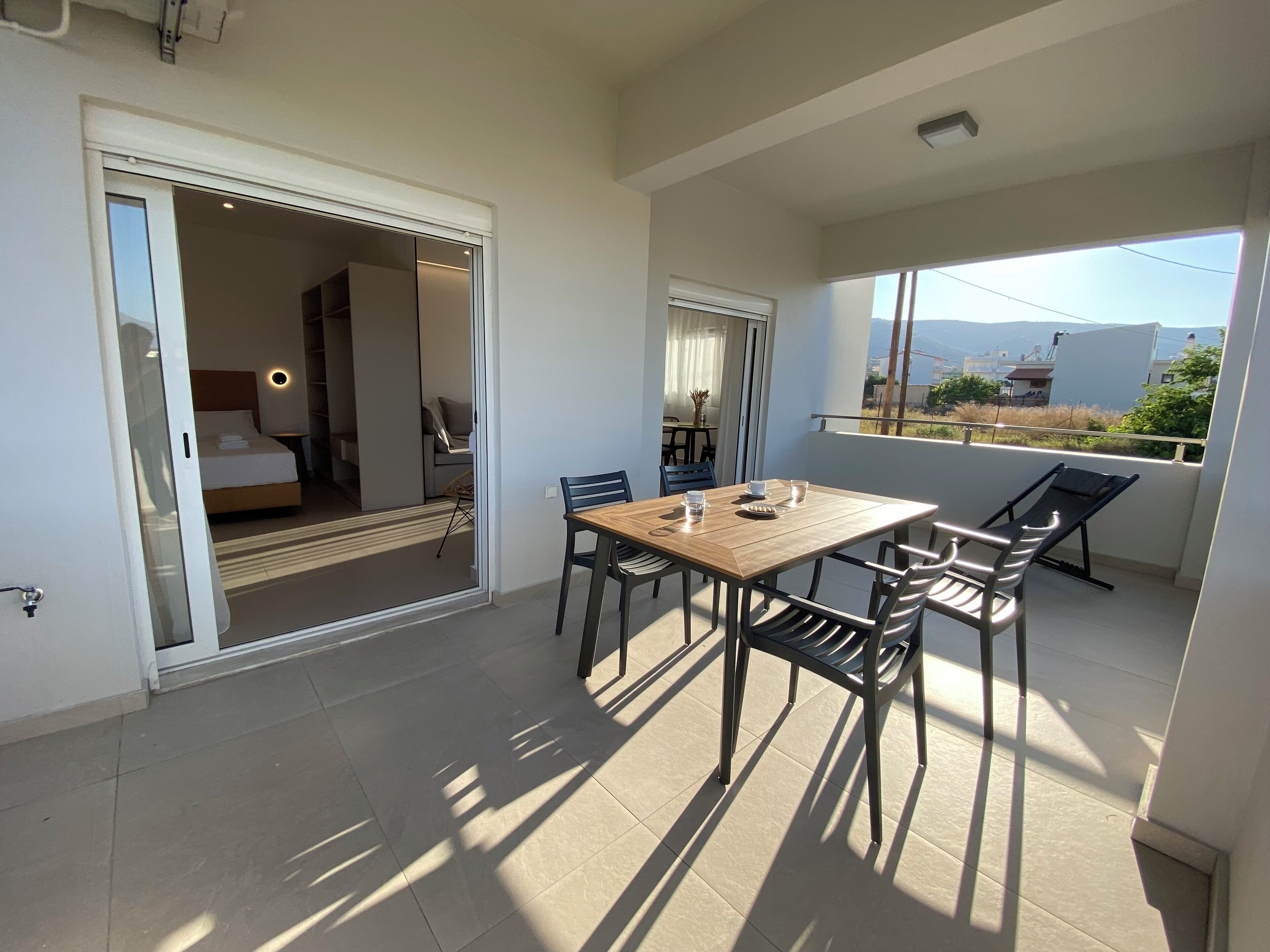 A balcony with a wooden table and chairs, a bed in the adjacent room, and a view of the landscape.