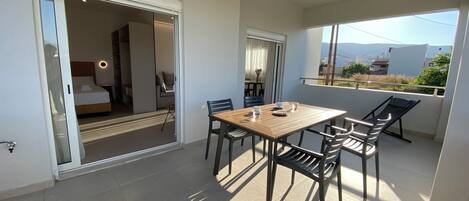 A balcony with a wooden table and chairs, a bed in the adjacent room, and a view of the landscape.