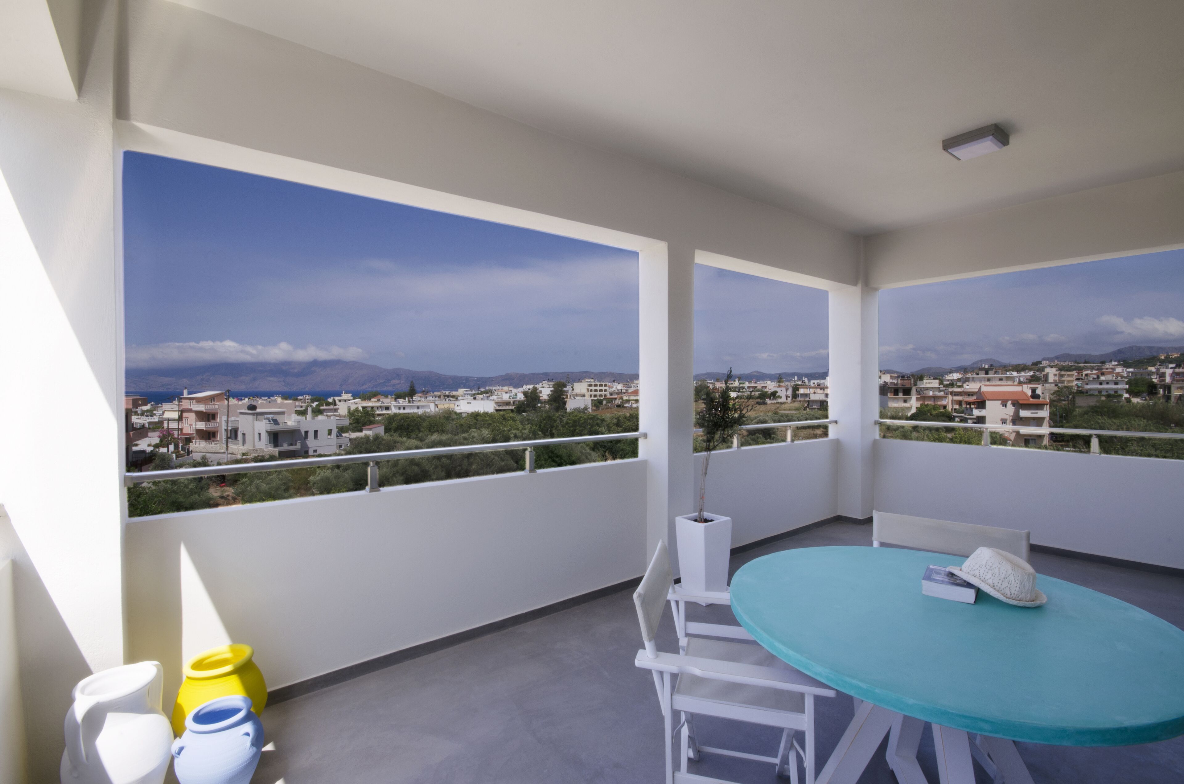 A balcony with a turquoise table and white chairs, overlooking a cityscape with mountains in the distance.