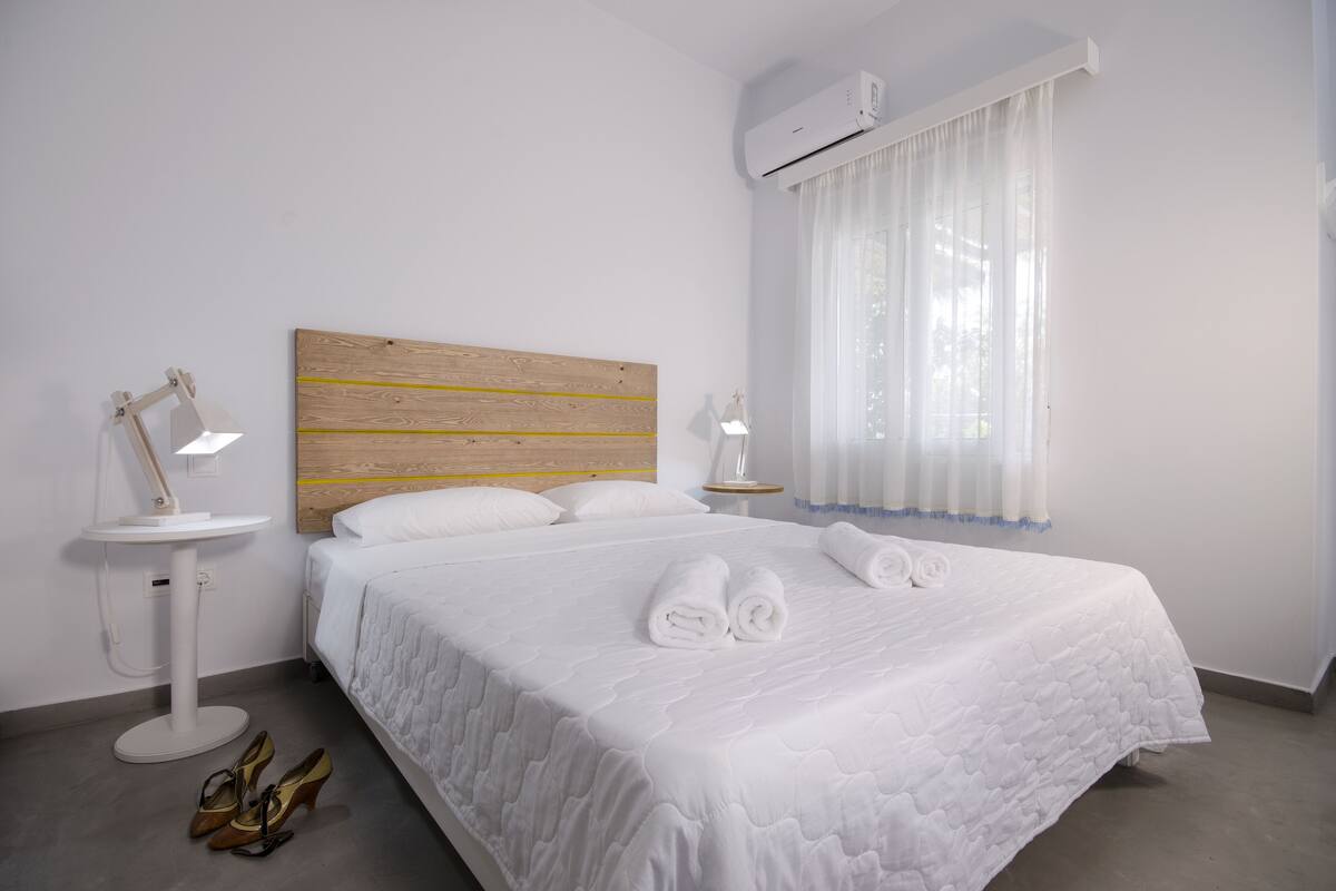 Una cama bien hecha con sábanas y toallas blancas, un cabecero de madera, una mesita de noche con lámpara y una ventana con cortinas traslúcidas.