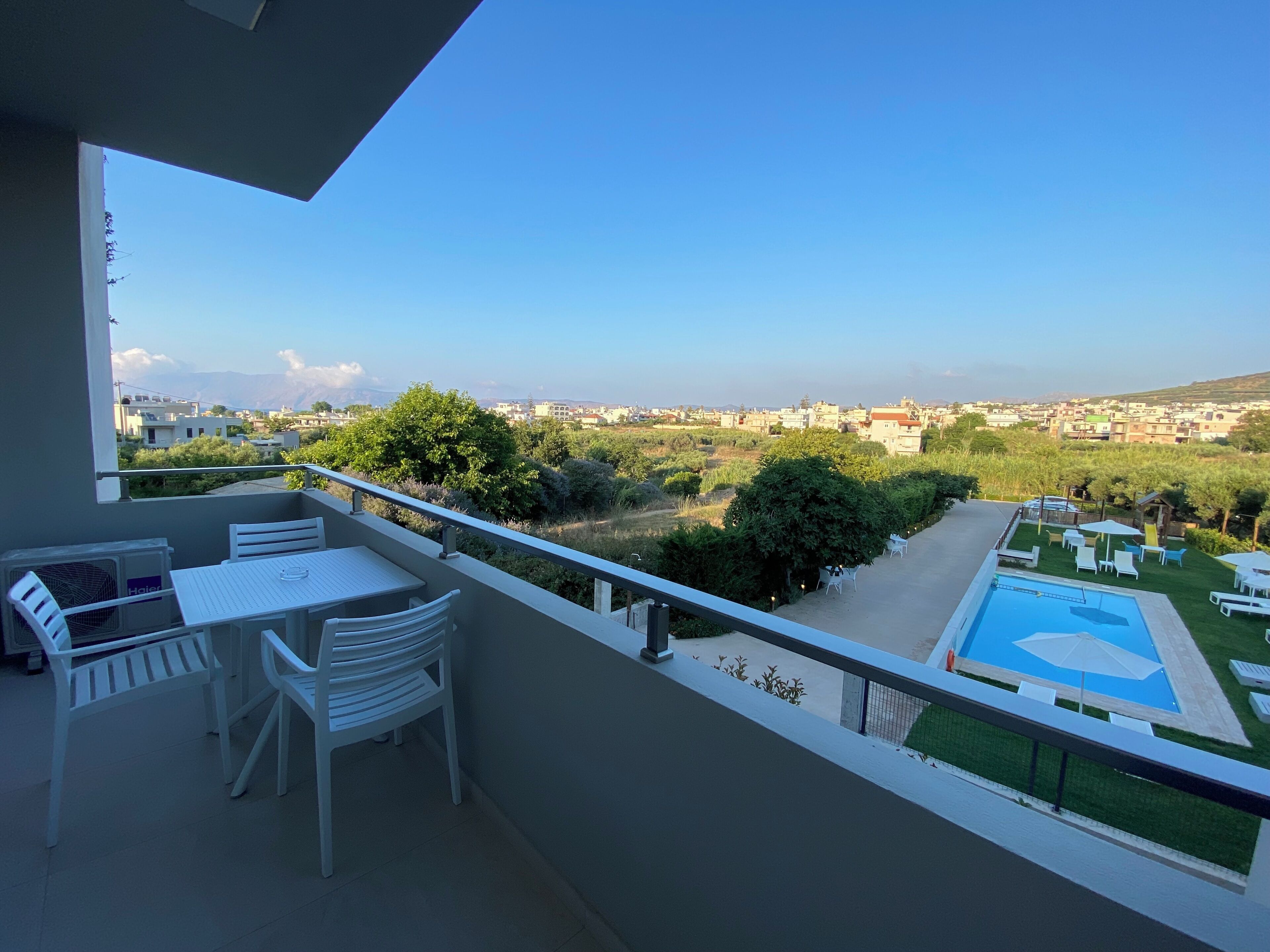 Ein Balkon mit Tisch und Stühlen, ein Poolbereich mit Liegestühlen und Blick auf eine Wohngegend mit Bäumen und Gebäuden.