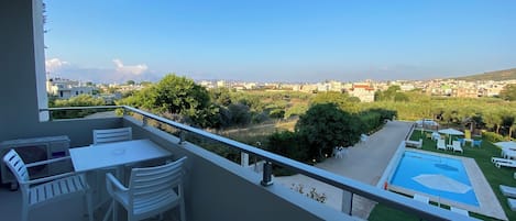 Ein Balkon mit Tisch und Stühlen, ein Poolbereich mit Liegestühlen und Blick auf eine Wohngegend mit Bäumen und Gebäuden.