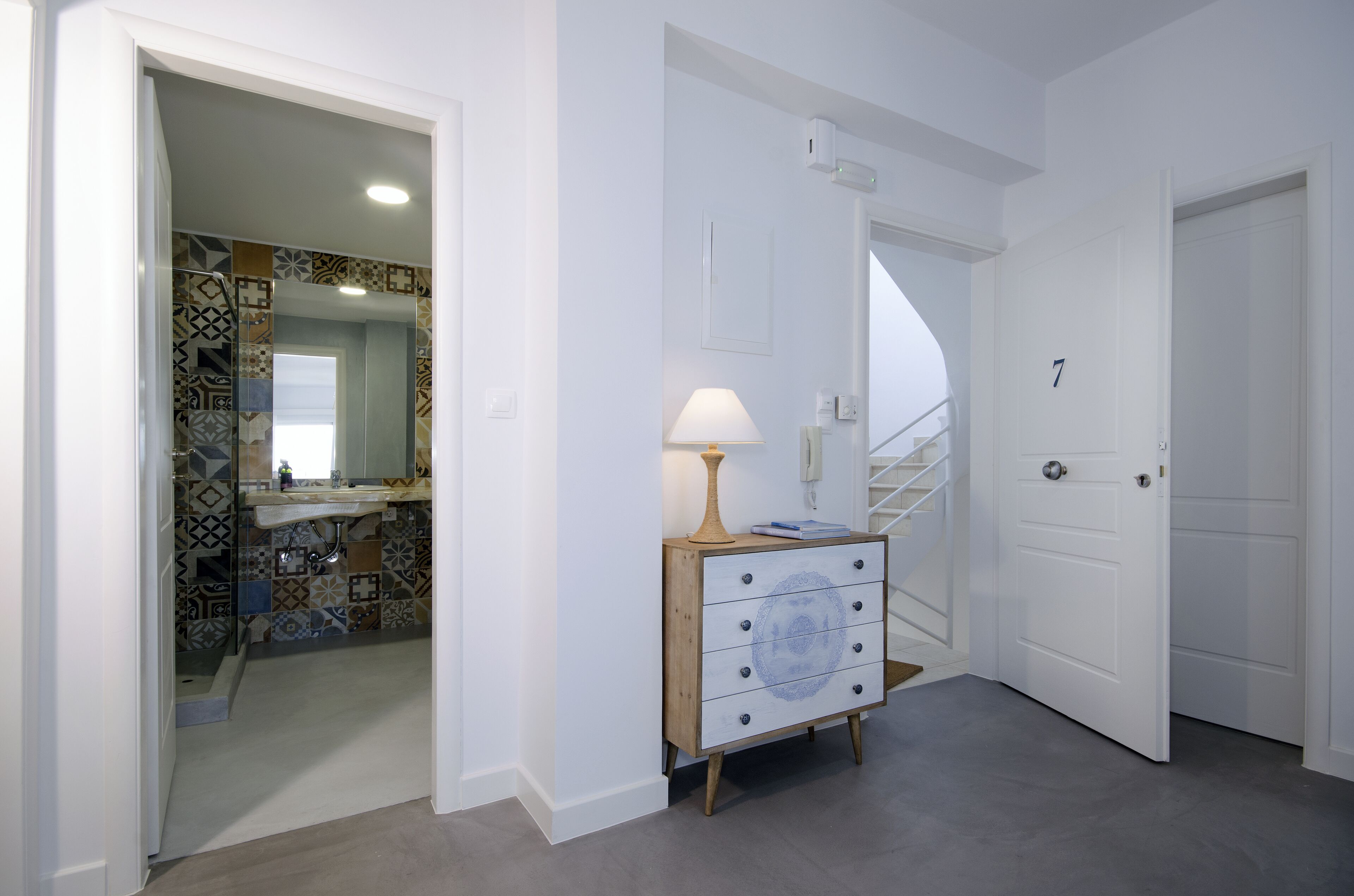 A hallway with a white door labeled '7', a wooden dresser with a lamp, and a bathroom with patterned tiles.