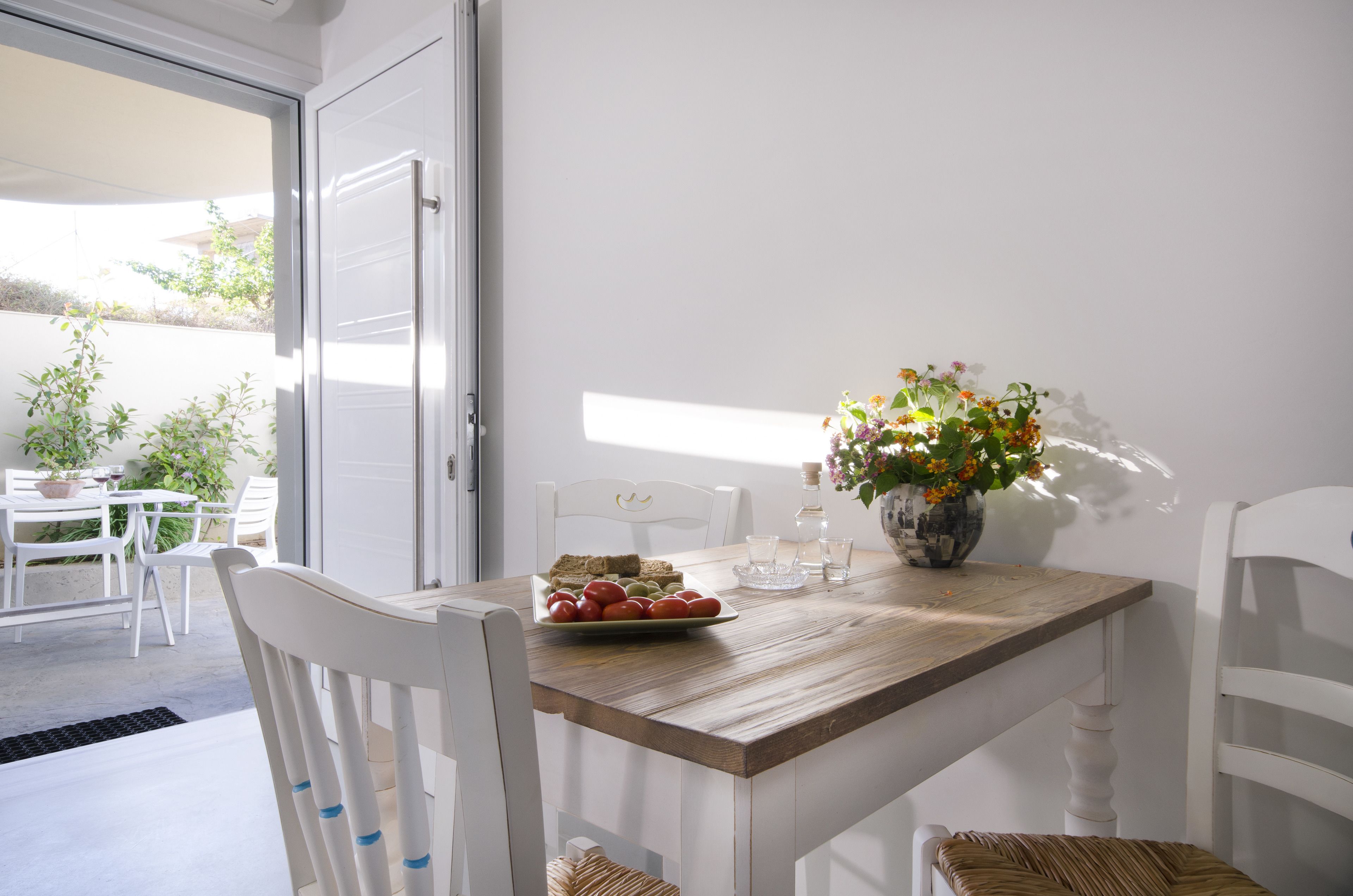 A dining area with a wooden table, white chairs, a vase with flowers, and a plate of tomatoes.