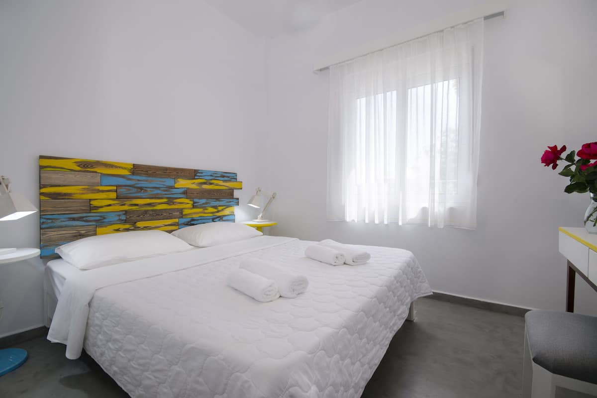 Una cama bien hecha con ropa de cama y toallas blancas, un cabecero de madera colorido, una mesita de noche con una lámpara y una ventana con cortinas ligeras.