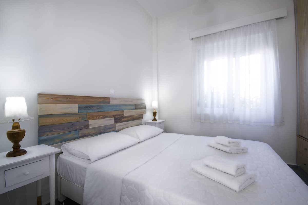 Una cama bien hecha con sábanas blancas y toallas dobladas, cabecera de madera, lámpara de noche y ventana con cortinas traslúcidas.