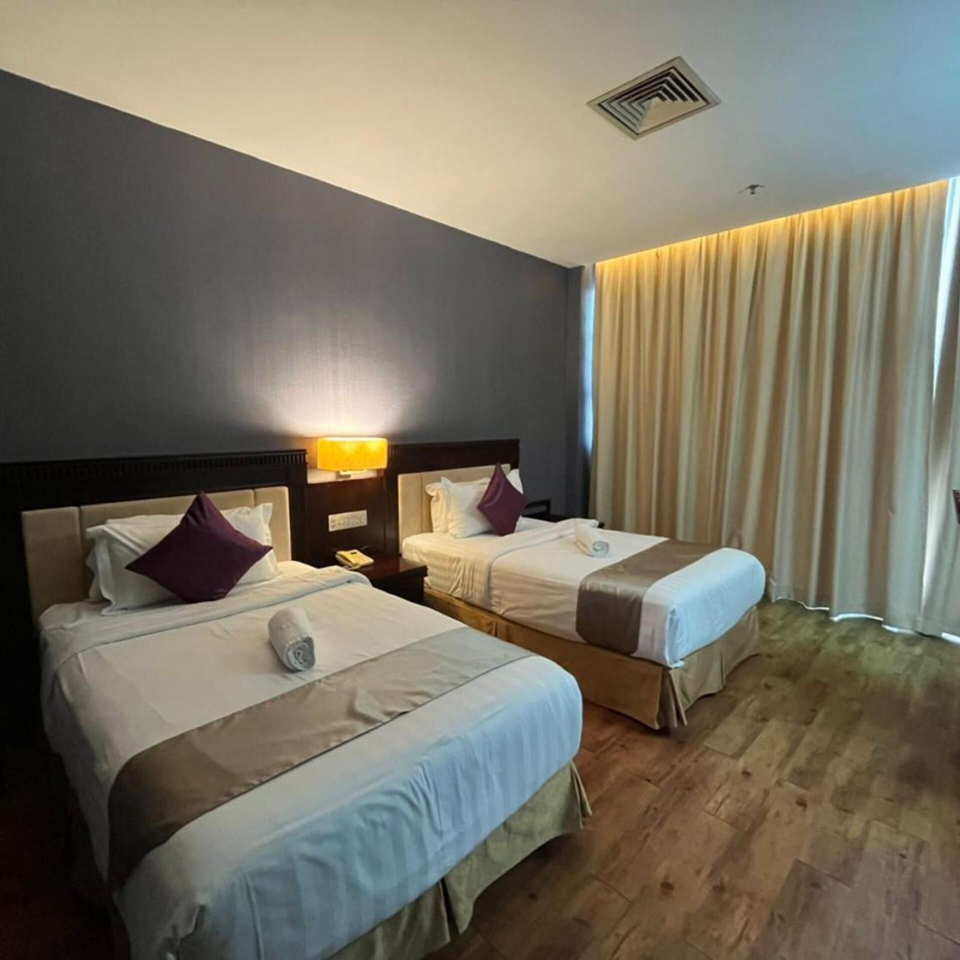 A hotel room with two beds, a nightstand with a lamp, a window with curtains, and a wooden floor.