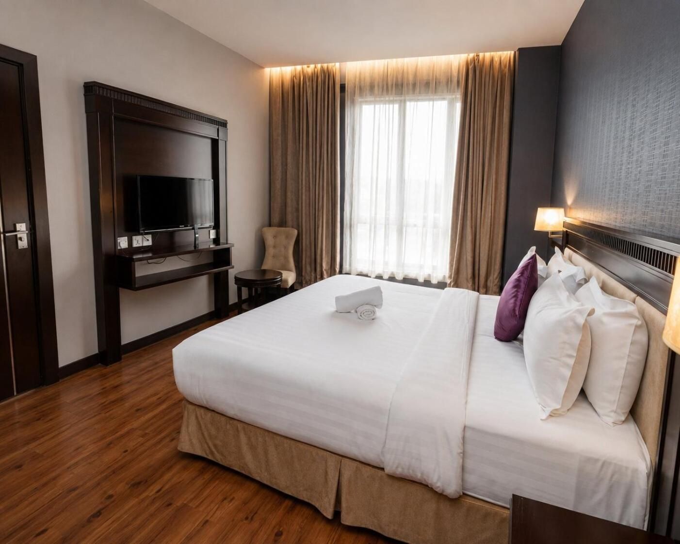 Habitación de hotel con una cama grande, un televisor montado en un mueble de madera, una silla y una ventana con cortinas.