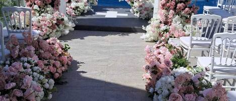 Ein schlichter Weg für eine Hochzeitszeremonie im Freien, geschmückt mit Blumen und weißen Stühlen, der zum Blick auf das Meer einlädt.