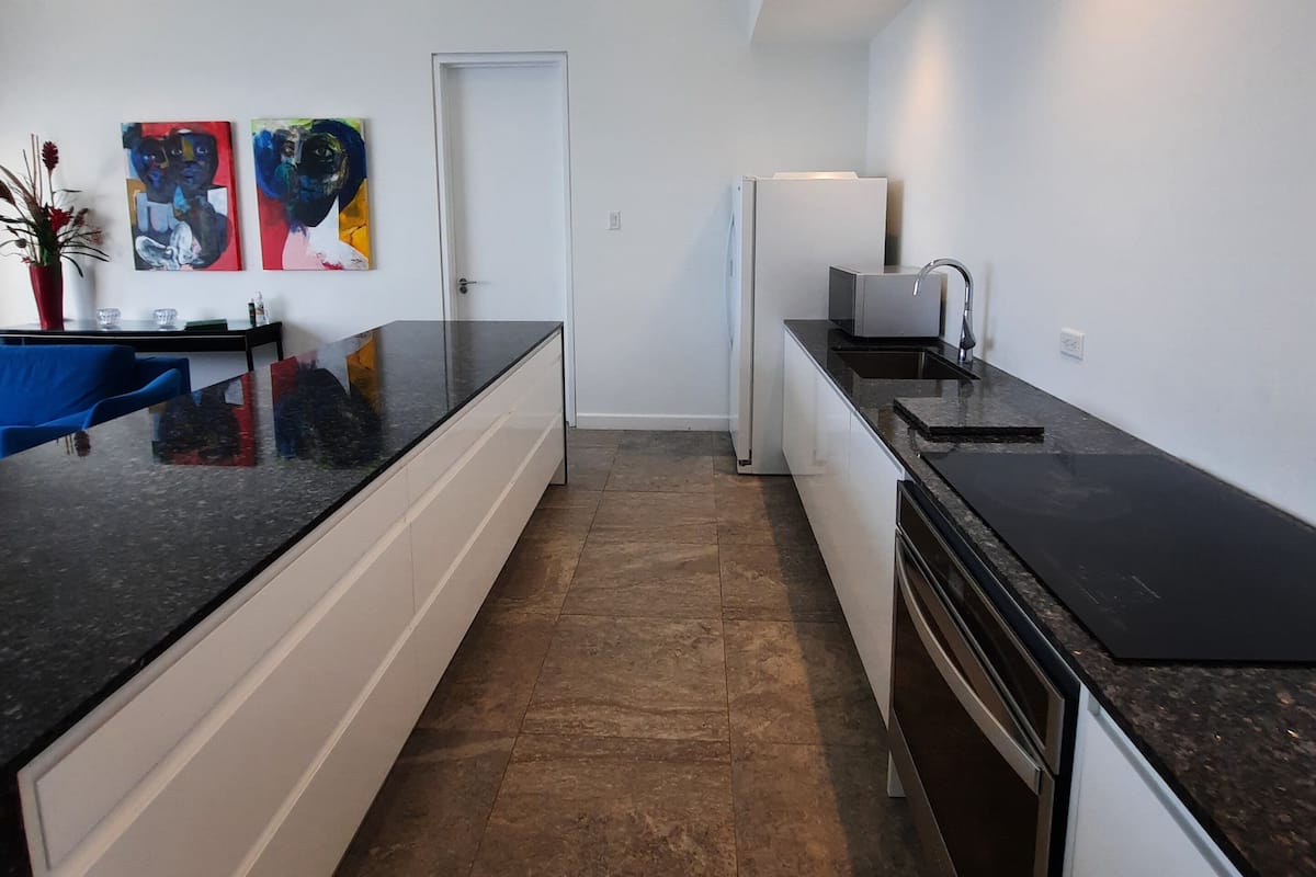 Een moderne keuken met witte kasten, zwarte aanrechtbladen en een betegelde vloer. Er hangen twee abstracte schilderijen aan de muur, en er is een deur en een koelkast.