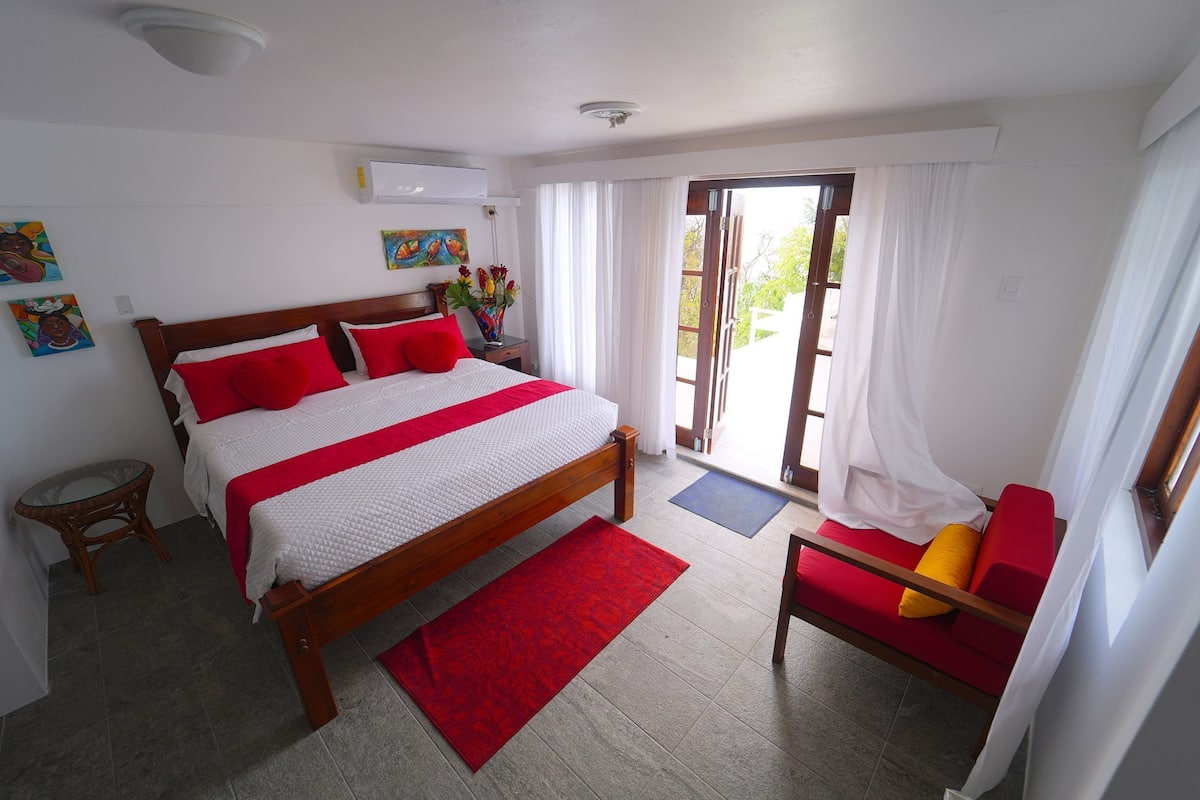 Een slaapkamer met een bed, een rode stoel, een tafeltje en een raam met gordijnen.