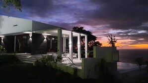Une maison moderne dotée d’un patio couvert et offrant une vue sur l’océan au coucher du soleil.