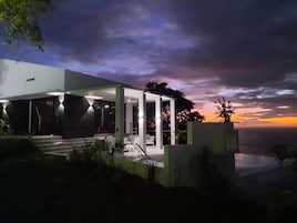 Uma casa moderna com varanda coberta e vista para o oceano ao pôr do sol.