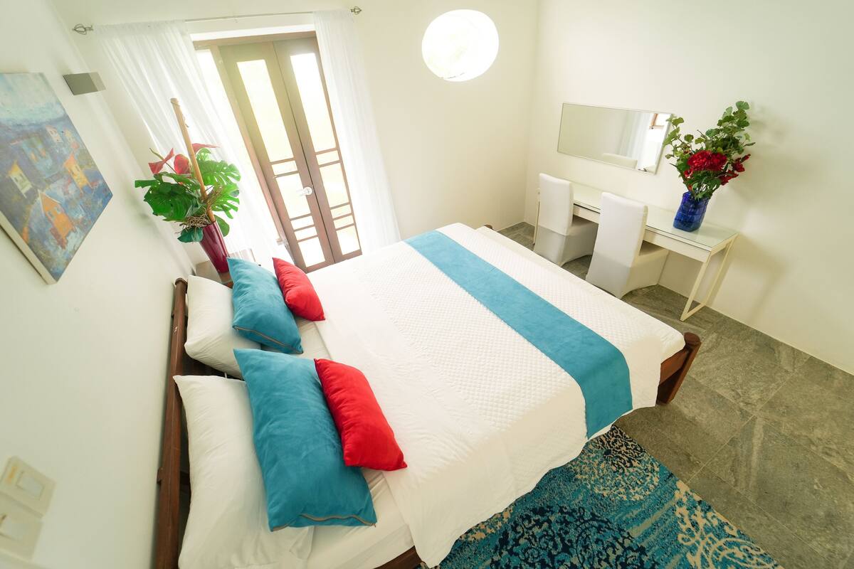 Een hotelkamer met een bed, een bureau, een stoel, een spiegel, een plant en een schilderij aan de muur.
