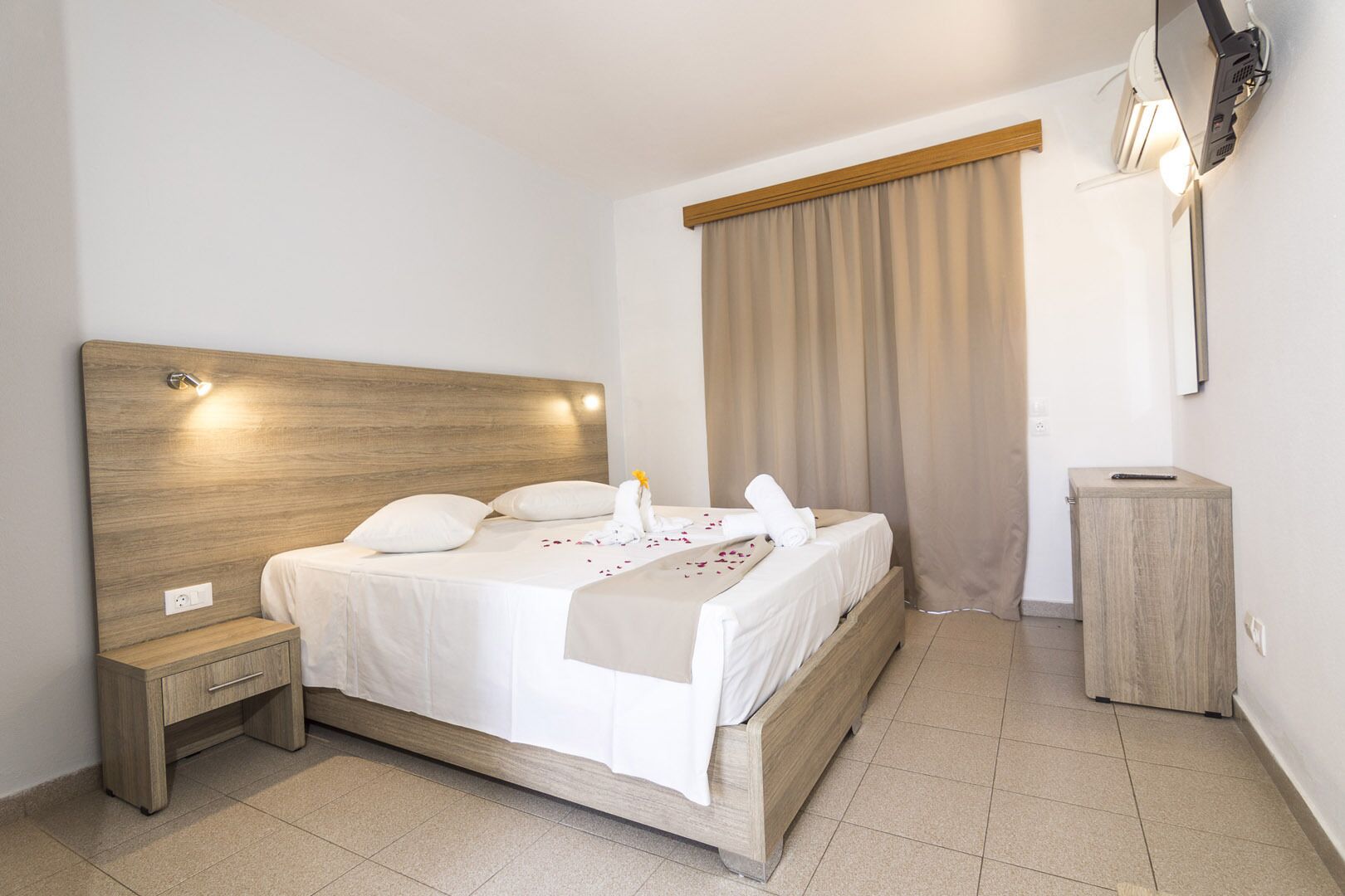 A neatly arranged hotel room with a wooden headboard, a bedside table, a small wooden cabinet, and a large window with curtains.