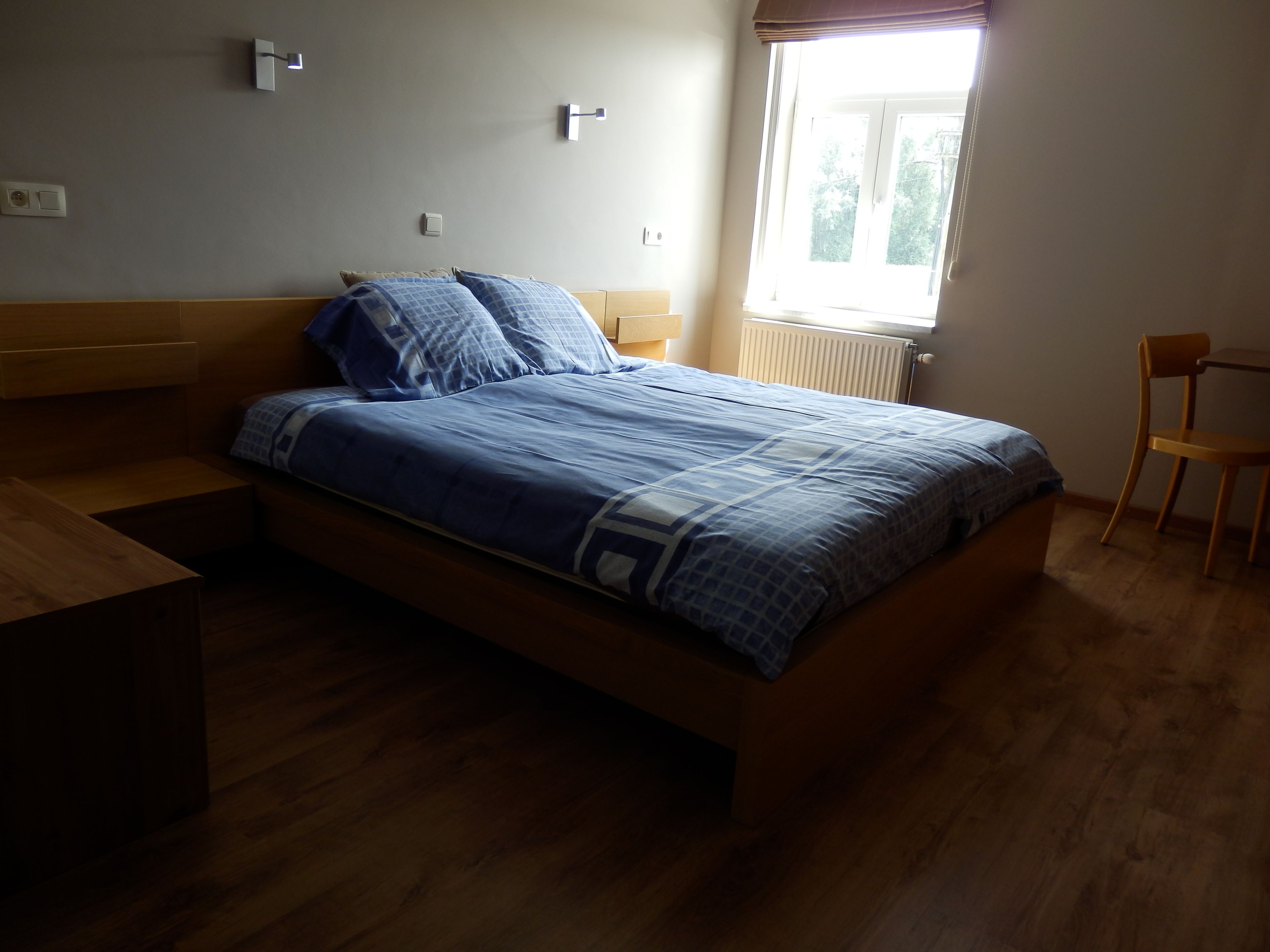 Ein Schlafzimmer mit einem hölzernen Bett, einem Fenster mit Jalousien, einem Stuhl und einer Heizung.