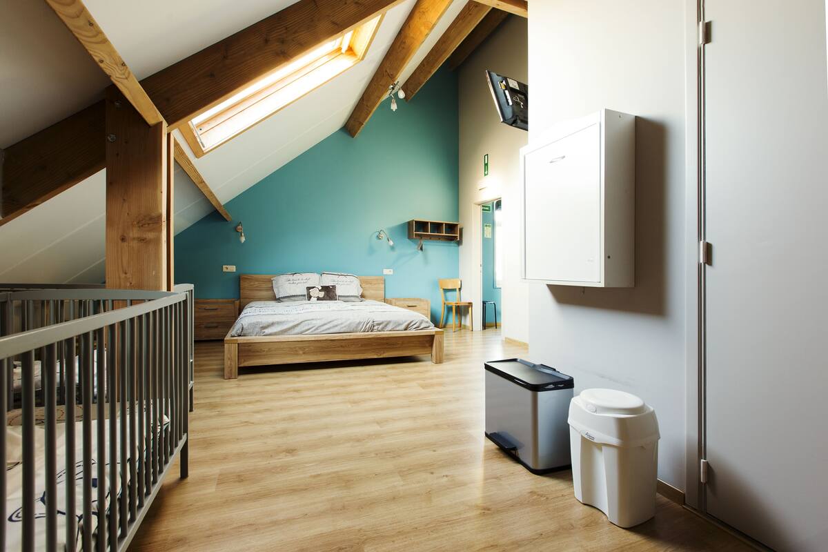 A bedroom with a wooden bed, a crib, a trash bin, and a TV mounted on the wall.