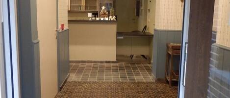A narrow hallway with patterned floor tiles, a kitchen area with cabinets and a sink, and a door leading to another room.