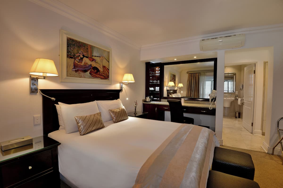 Quarto de hotel com uma cama grande, duas luminárias de cabeceira, um quadro na parede, uma escrivaninha com um espelho e um banheiro visível através de uma porta aberta.