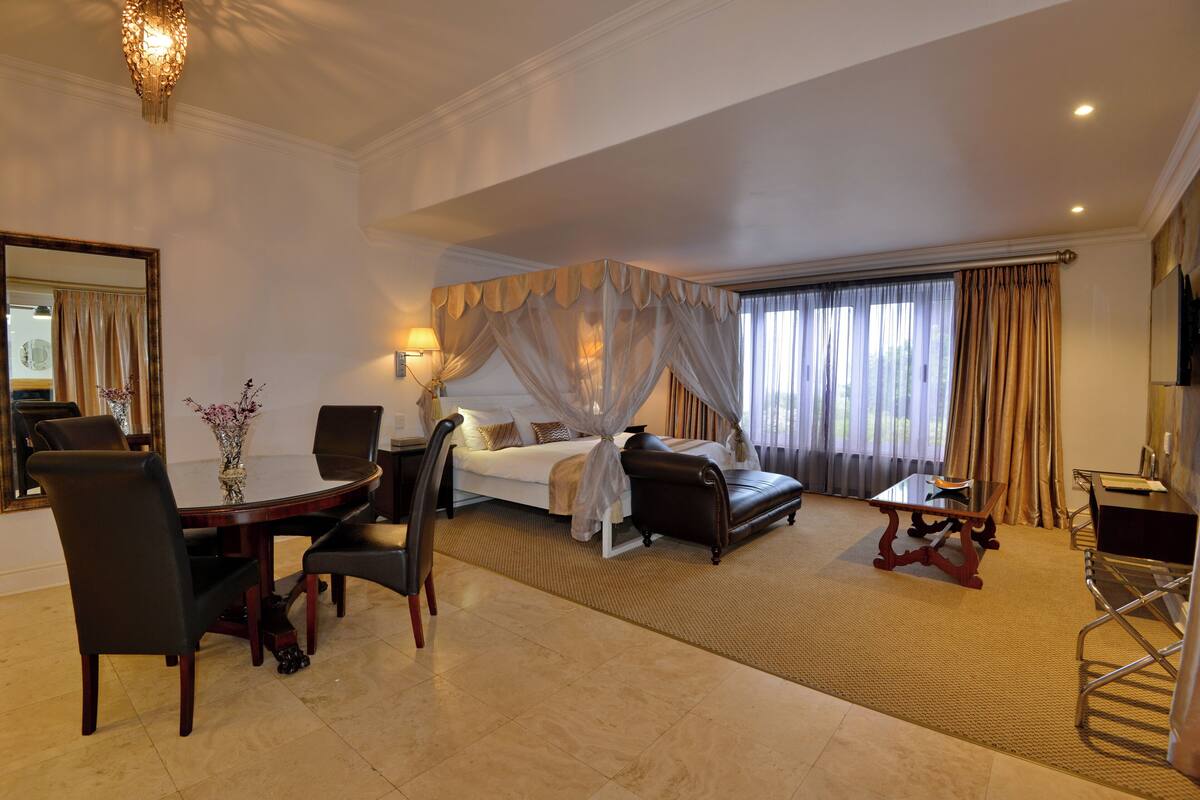Quarto de hotel com cama, mesa de jantar com cadeiras, pequena mesa com uma cadeira, espelho grande e janela com cortinas.