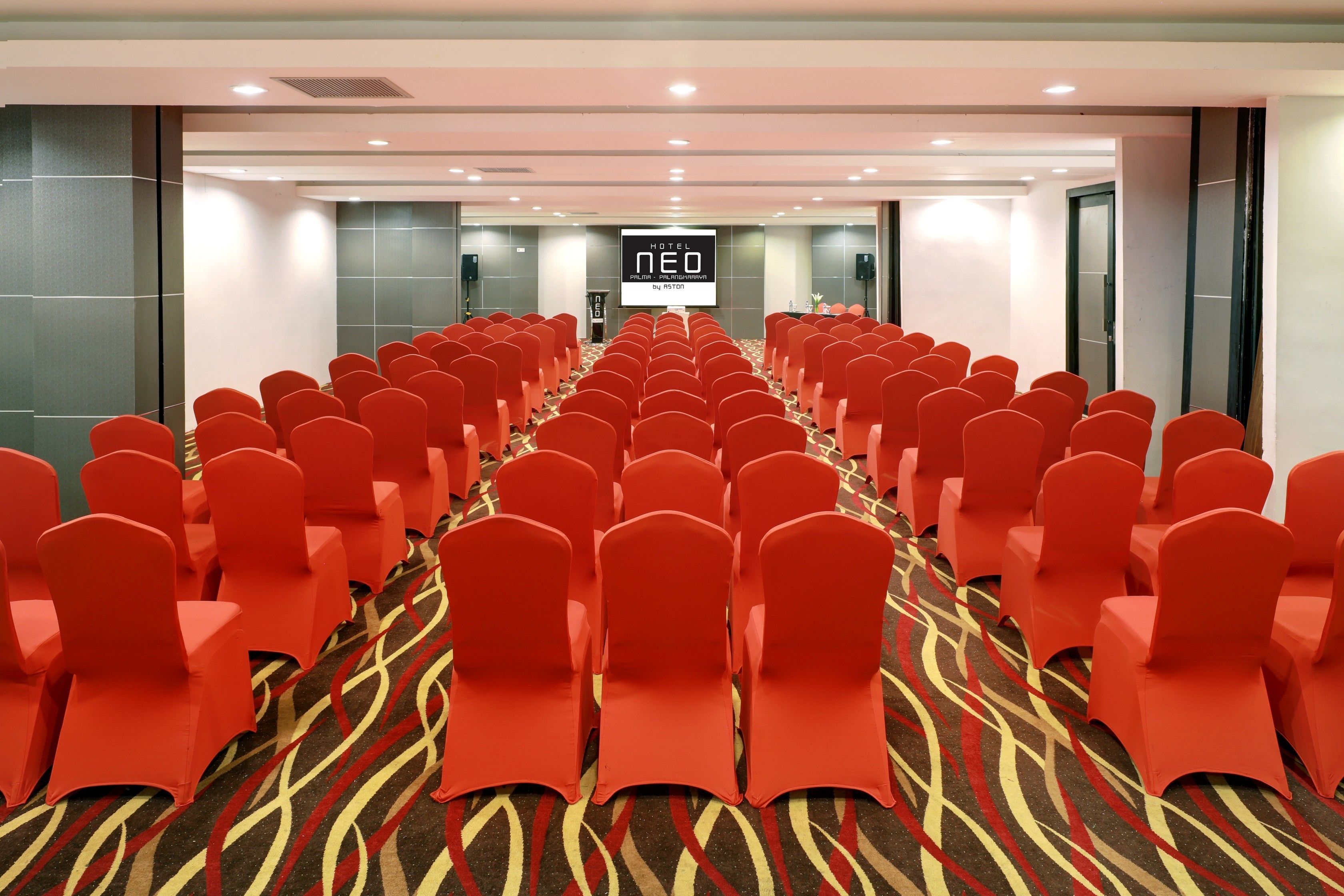A hotel conference room with rows of red chairs and a carpeted floor.