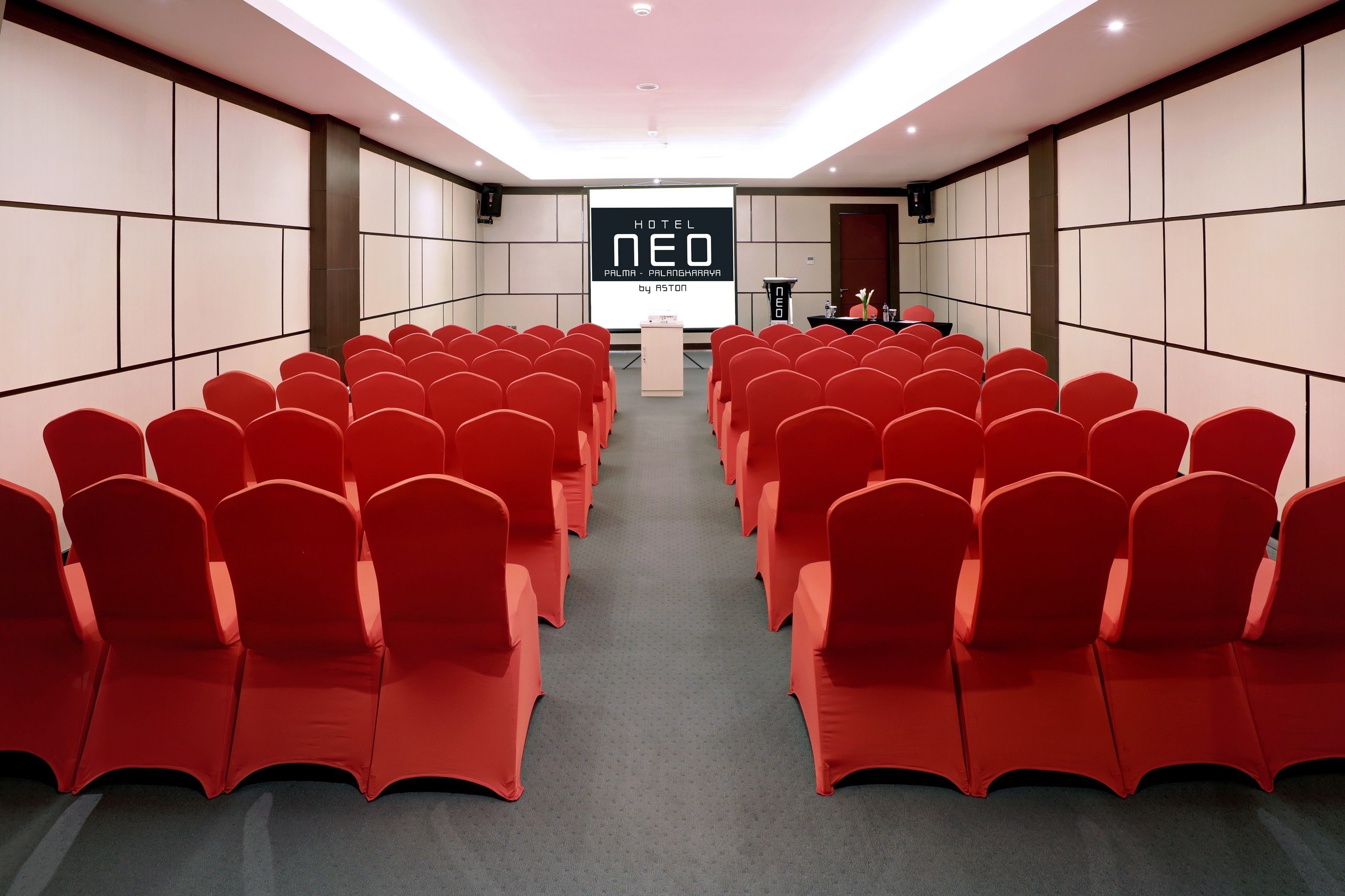 A conference room with rows of red chairs, a podium, and a screen displaying 'HOTEL NEO PALANGBARAWA by ASTON'.