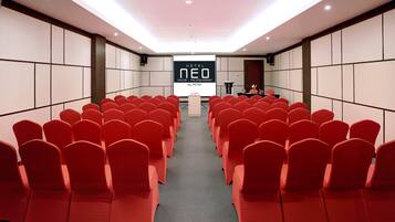 A conference room with rows of red chairs, a podium, and a screen displaying 'HOTEL NEO PALANGBARAWA by ASTON'.