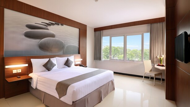 A modern hotel room with a large bed, a flat-screen TV, a desk with a chair, and a window with a view of trees. - Hotel Neo Palma - Palangkaraya by ASTON (Palangkaraya)