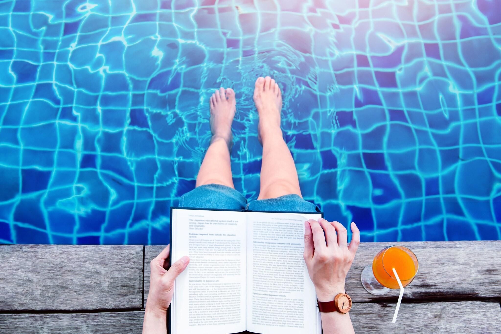 A person relaxing by a pool with a book and a drink.