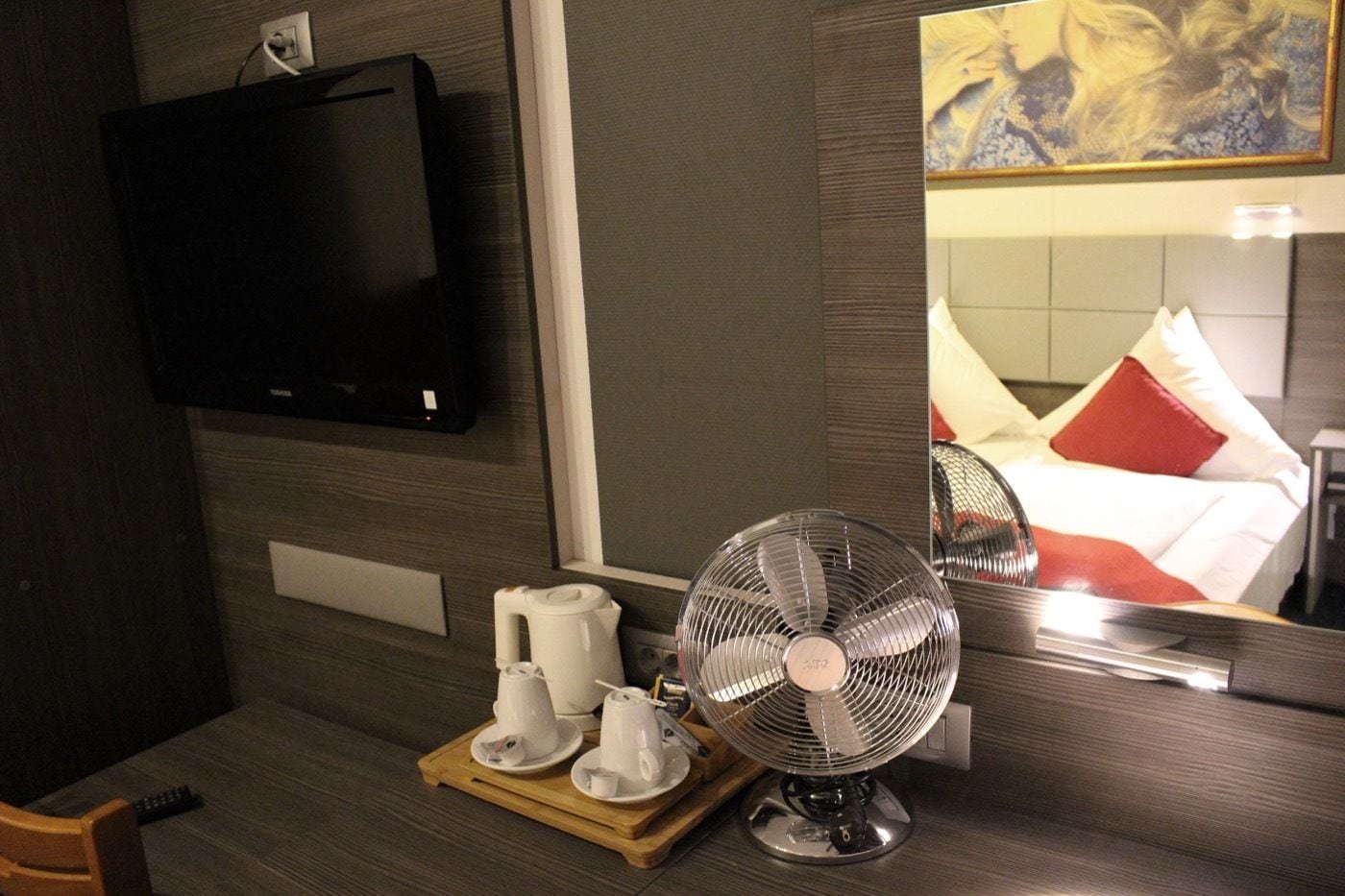 A hotel room with a TV mounted on the wall, a bedside table with a fan, a tray with cups and a kettle, and a bed with red and white bedding.