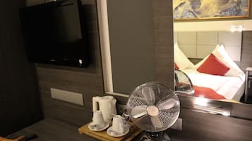 Una habitación de hotel con un televisor montado en la pared, una mesita de noche con un ventilador, una bandeja con tazas y una tetera, y una cama con ropa de cama roja y blanca.