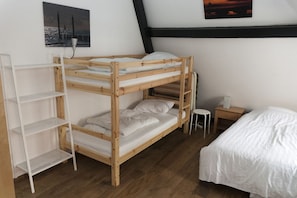 A bunk bed with a white mattress, a white shelf unit, a small white stool, and a wooden nightstand with a white lamp. - City Hostel Vlissingen (Vlissingen)
