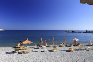 A beach with pebbles, umbrellas, and lounge chairs. - Maria Studios and Apartments (Rhodes)